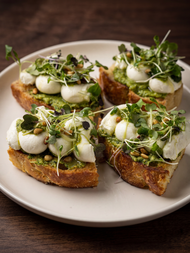 Spring onion pesto focaccia toast with whipped goat cheese, pine nuts, and spring greens on a white plate