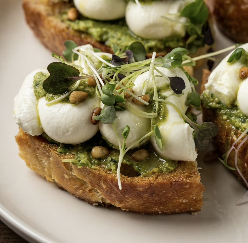 Spring onion pesto focaccia toast with whipped goat cheese, pine nuts, and microgreens on a white plate